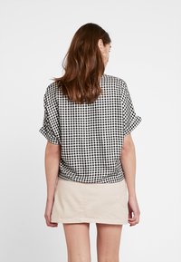 En kvinna står med ryggen mot kameran, iklädd en svartvit rutig skjorta och en ljusbeige kjol mot en vit bakgrund.