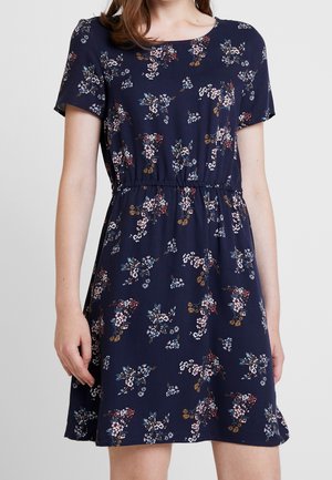 Frau trägt ein marineblaues Kleid mit kurzen Ärmeln, einem gerafften Taillenbereich und einem kleinen, mehrfarbigen Blumenmuster.