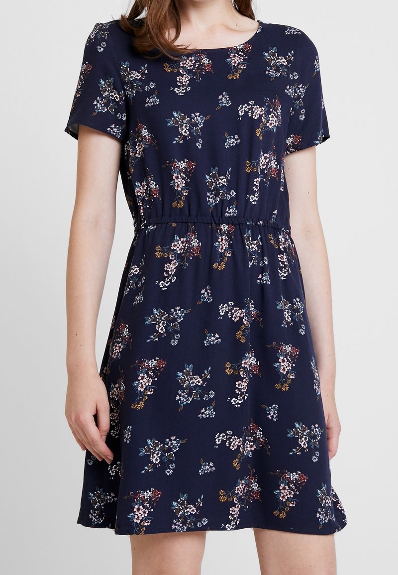 Kobieta w granatowej sukience z krótkim rękawem, z marszczonym stanem i małym, wielokolorowym kwiatowym wzorem.