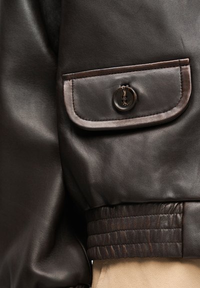 Μανίκι σκούρας καφέ δερμάτινου μπουφάν με τσεπάκι με καπάκι και κουμπί και ριμπ μανσέτα πάνω σε μπεζ υφασμάτινο φόντο.