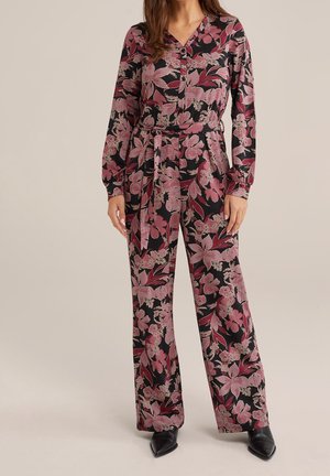 Frau trägt einen schwarzen Overall mit rosa Blumenmuster, langen Ärmeln, geknöpftem Vorderteil, Gürtel in der Taille und weit geschnittenen Hosen, kombiniert mit schwarzen Schuhen.