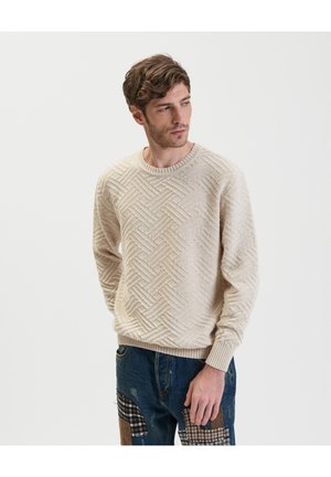 Beżowy sweter dzianinowy z teksturowanym wzorem w jodełkę, okrągłym dekoltem i ściągaczami na rękawach. W połączeniu z niebieskimi dżinsami w stylu patchwork.