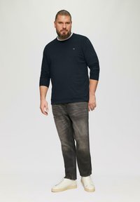 Marineblaue Langarmbluse mit Rundhalsausschnitt, kombiniert mit verwaschenen grauen Jeans. Weiße Sneaker mit strukturierten Details und einer cremefarbenen Gummisohle.