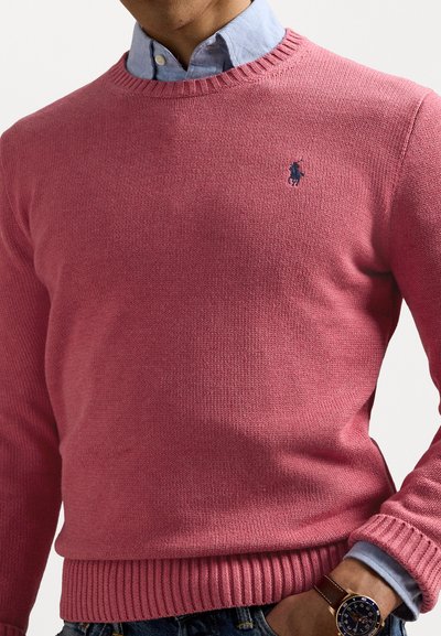 Rožinis megztinis su briaunotais rankogaliais ir apdaila, ant krūtinės esantis mažas mėlynas logotipas. Dėvimas virš šviesiai mėlynos pusiau apykaklės marškinėlių.