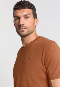 Bruin T-shirt met korte mouwen, gemaakt van zachte stof, met een ronde halslijn en een klein logo aan de linkerkant van de borst.