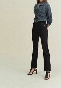 Jean noir à jambe droite associé à une chemise en denim foncé avec deux poches poitrine, et des sandales noires à talon bloc avec un bout ouvert.