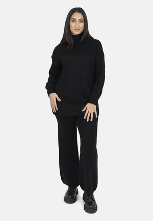Schwarzer gestrickter Pullover mit hohem Kragen und weiten Ärmeln, kombiniert mit passenden weiten Hosen. Schwarze klobige Schuhe runden das Outfit ab.