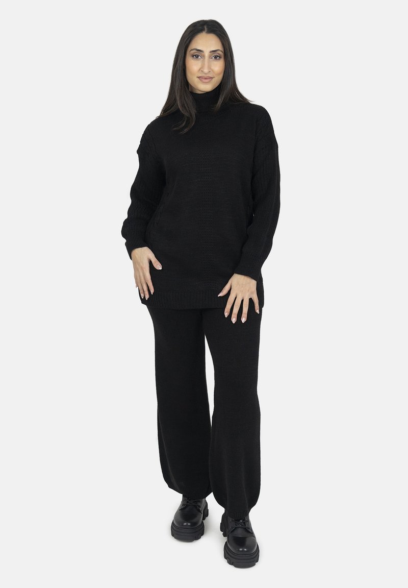 Maglione nero lavorato a maglia con collo alto e maniche larghe, abbinato a pantaloni a gamba larga coordinati. Scarpe chunky nere completano l'outfit.