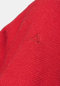 Tissu rouge texturé avec un petit logo triangulaire brodé dans un fil rouge assorti.