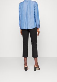 Camisa de mezclilla azul claro con detalle de botones en la espalda, combinada con pantalones negros recortados y sandalias negras de tacón.
