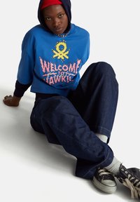 Modrá zkrácená tričko s grafikou "VÍTEJTE V HAWKINS," v kombinaci s tmavými džínami, šedými ponožkami a černými teniskami. Viditelná těsně přiléhající kapuce.