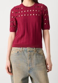 Lyhyet hihat, ribattu neulepusero viininpunaisena, jossa on pyöreitä aukkoja harteilla. Yhdistetty vaaleansinisiin, korkeavyötäröisiin farkkuihin.