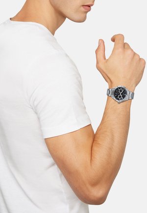 Montre en acier inoxydable avec un cadran noir et des accents argentés, positionnée sur un poignet, partiellement visible avec un t-shirt blanc.