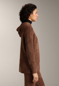 Žena s kučeravými vlasmi oblečená v hnedej textúrovanej mikine s kapucňou a zodpovedajúcich nohaviciach, stojaca bokom na jednoliatom svetlom pozadí.