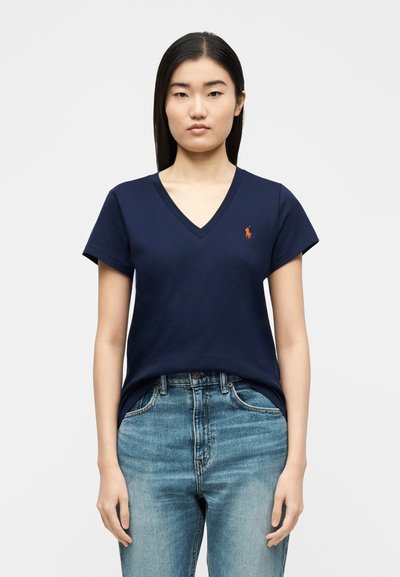 Νεαρή γυναίκα με ίσια σκούρα μαλλιά, φοράει μπλε μπλούζα V με μικρό πορτοκαλί λογότυπο και μπλε τζιν, στέκεται μπροστά σε απλό φόντο.