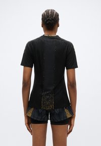 Črna športna majica z teksturirano tkanino, z zlatimi poudarki in oprijeto zasnovo. Kombinirana s črnimi kratkimi hlačami z vzorcem in volančastim robom.