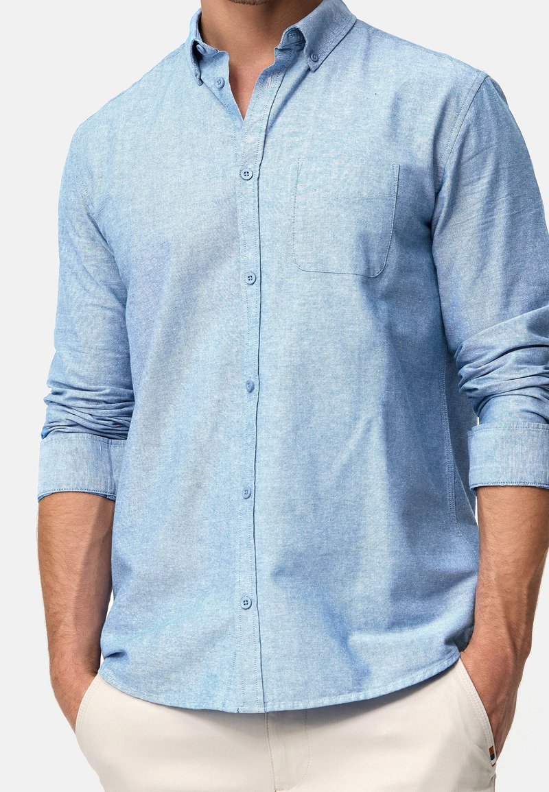 Homme portant une chemise bleu clair à boutons avec manches retroussées et pantalon beige, mains dans les poches, sur un fond uni.