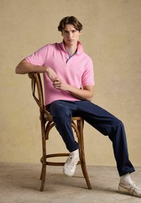 Jeune homme en polo rose et pantalon bleu marine assis de côté sur une chaise en bois devant un fond beige.