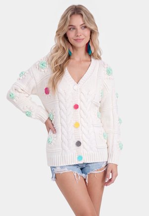 Jonge vrouw draagt een witte gebreide cardigan met kleurrijke pomponknopen en bloemdetaillering, gecombineerd met gescheurde denim shorts.