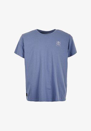 Jong persoon met krullend haar, gekleed in een blauw T-shirt en zwarte broek, staand buiten bij een reling met zichtbare tatoeages op de armen.