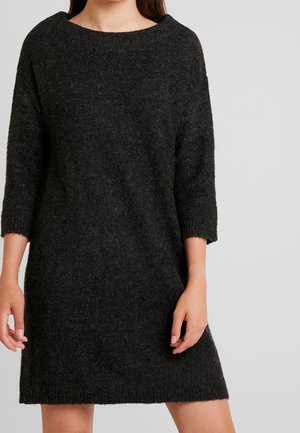 Kvinde iført en mørkegrå, langærmet, strikket sweaterkjole, der står mod en ensfarvet lys grå baggrund.