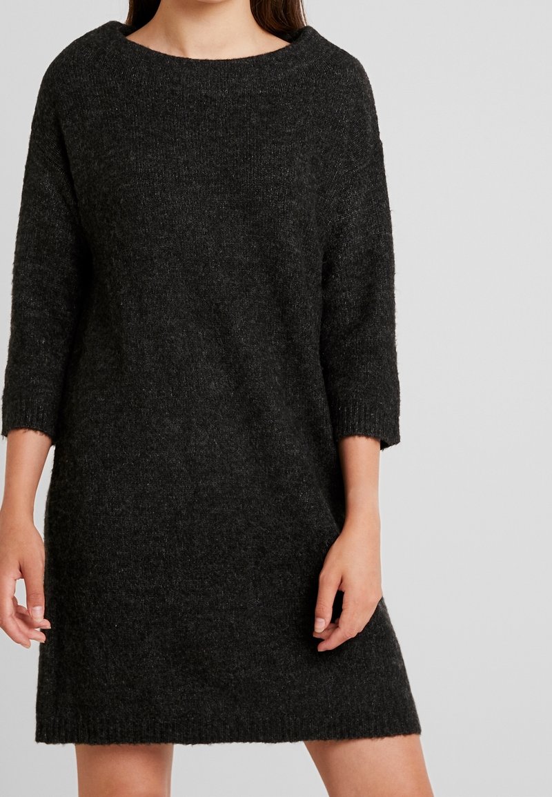 Femme portant une robe pull en tricot gris foncé à manches longues, debout devant un fond uni gris clair.