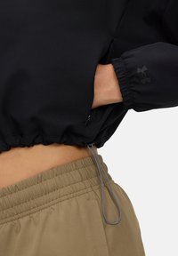 Chaqueta negra corta con dobladillo fruncido, que presenta un bolsillo lateral y puños elásticos. Combinada con pantalones de cintura elástica color beige.