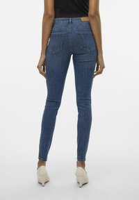 Mėlynos džinsinės liesos kelnės, pasiūtos iš džinsinio audinio, turinčios prigludusią siluetą, su dviem galinėmis kišenėmis ir ruda odine etikete ant juostos.