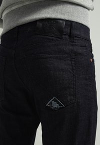 Jeans in denim di un blu scuro con una finitura testurizzata, dotati di un'etichetta in pelle nera, una toppa a forma di triangolo e dettagli in rivetti sulla vita.