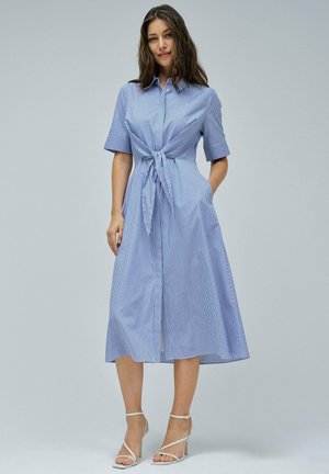 Robe chemise à rayures bleues et blanches avec des manches courtes, taille cintrée et ourlet au genou. Le mannequin porte des talons à brides.