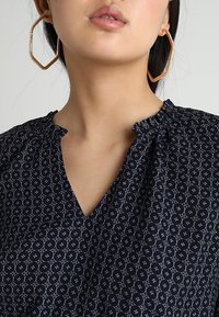 Navy blouse with circular white patterns, ruffled neckline detail, and a V-shaped opening. Large geometric earrings in light metal.