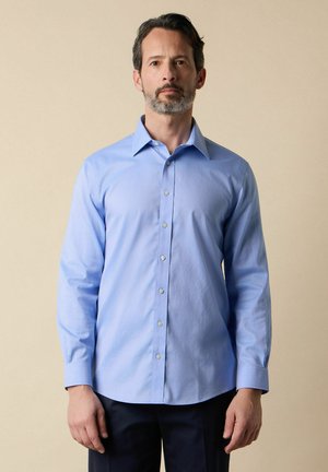 Mann mit dunklem Haar und Bart, der ein hellblaues langärmliges Hemd mit Knöpfen und dunkle Hose trägt, vor einem schlichten beigen Hintergrund stehend.