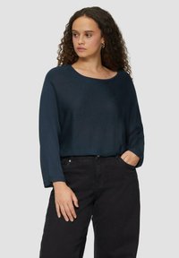 Dunkelblauer Strickpullover mit weitem Ausschnitt und dreiviertel Ärmel, kombiniert mit schwarzen hoch geschnittenen Hosen, Hände in den Taschen.