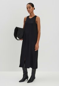 Schwarzes ärmelloses Kleid mit gerader Silhouette, seitlichem Schlitz, getragen mit knielangen schwarzen Stiefeln und einer strukturierten schwarzen Handtasche.