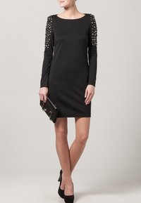 Robe noire à longues manches avec une silhouette ajustée, ornée d'accents cloutés sur les épaules. Le matériau est lisse et extensible. Elle tient une petite pochette noire.