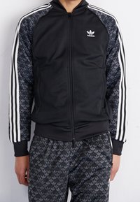 Černá bunda na zip s šedými rukávy se vzorem a bílými pruhy po stranách. Kombinováno se stejnými černými kalhotami s logem vzoru.