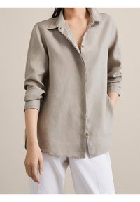 Chemise en lin gris clair avec col, boutons sur le devant, manches longues retroussées et poches latérales ; texture lisse et coupe décontractée.