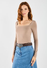 Långärmad topp med fyrkantig halsringning i ljusbrunt tyg, tillsammans med en högmidjad denimkjol. Outfiten har ett flätat brunt bälte med en silverspänne.