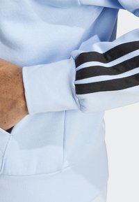 Ljusblå sweatshirt med svart tre-stripes-design på ärmarna, ribbade muddar och en texturerad tyg för ökad komfort.