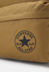 Borsa a spalla in tela color cannella con un logo rotondo nero "Converse All Star" con dettagli a stella e testo. Texture liscia e design resistente.