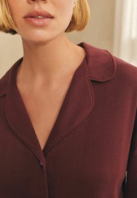Primo piano di una persona con capelli biondi corti che indossa una camicia colletto bordeaux texture con bottoni davanti.