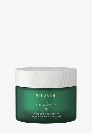 Tarro de vidrio verde oscuro con una tapa de madera blanca texturizada. Texto dorado etiqueta el producto como "RITUALS THE RITUAL OF JING crema corporal batida."