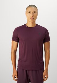 Homme portant un t-shirt de sport cintré à manches courtes bordeaux et un short assorti, se tenant devant un fond blanc uni.