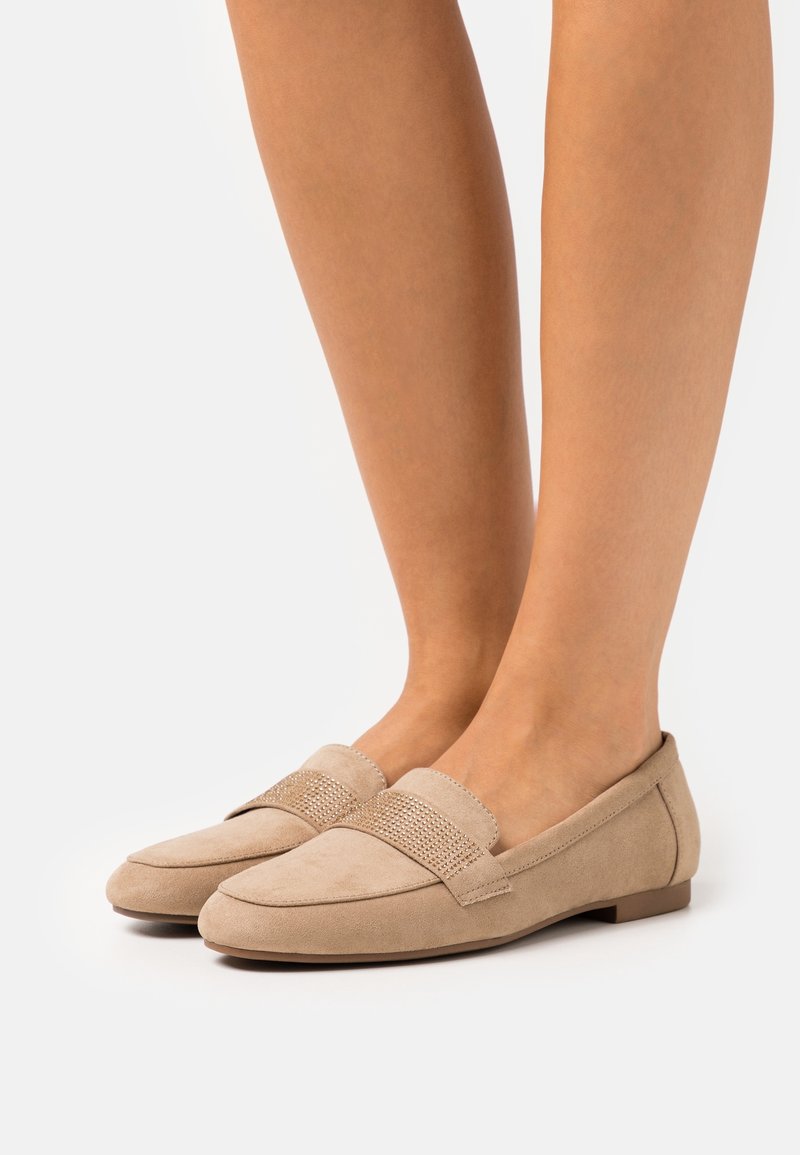 Mocassins en daim beige avec un petit talon et une bande texturée, portés pieds nus sur fond blanc.