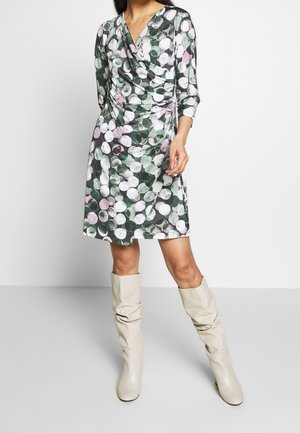 Kobieta w sukience do kolan z długim rękawem, w zielono-biało-różowy wzór w kółka, w połączeniu z białymi, luźnymi kozakami sięgającymi kolan.