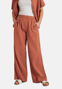 Brede, roestkleurige broek van textuurstof met een elastische tailleband en zijzakken; gecombineerd met een witte top en sandalen.