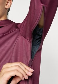 Hand opening zipper under armpit of purple jacket, revealing black inner layer, against a plain light background.