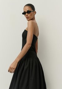 Čierne šaty bez ramienok s priliehavým korzážom a objemnou sukňou so zhromaždením. Modelka má na sebe elegantné úzke čierne slnečné okuliare.