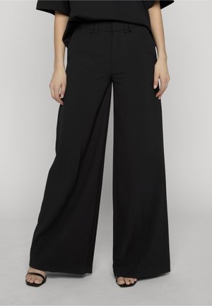 Femme portant un pantalon noir à jambes larges et des sandales noires à bouts ouverts, debout devant un fond clair uni.