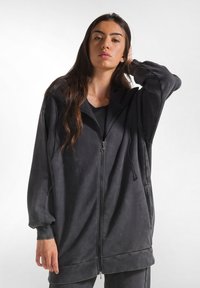 Felpa nera lunga con zip, realizzata in tessuto morbido e dal fit rilassato, con cappuccio regolabile e tasche laterali.